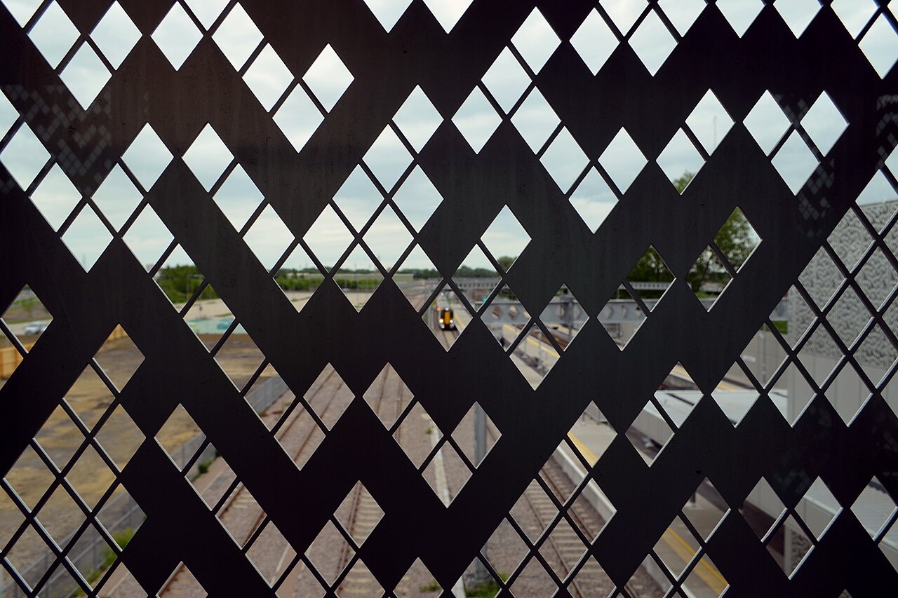 Looking through the perforated aluminum cladding of Cambridge North railway station: bright diamond-shaped holes in a Rule 30 pattern, with a train arriving on the tracks beyond